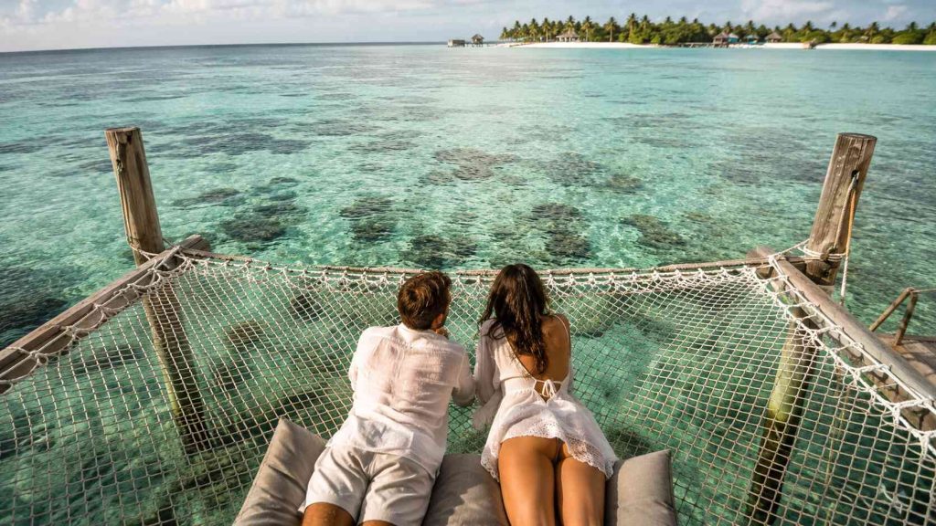Couple on suspended hammock net above crystal clear turquoise Maldives lagoon at Gili Lankanfushi with coral reef visible below